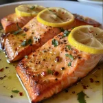 Salmon fillet glistening with lemon butter sauce alongside fresh lemon slices and a sprinkle of parsley.