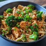 Close-up shot of a hearty noodle bowl with spicy garlic chicken, broccoli florets, and a drizzle of sauce.