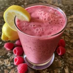 Close-up view of a vibrant raspberry lemon smoothie in a glass, with fresh raspberries and lemon slices beside it.