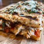 A close-up view of a grilled chicken parmesan flatbread sandwich, showcasing layers of chicken, cheese, and roasted tomato jam.