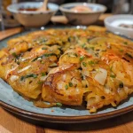 ['Close-up view of a juicy Haemul Pajeon, showcasing seafood and green onions.', 'Side view of a crispy Korean seafood pancake, garnished with scallions and chili.', 'Juicy Haemul Pajeon with a golden brown crust, highlighting mixed seafood ingredients.', 'Detailed image of Korean seafood pancake, featuring shrimp, squid, and sliced scallions.']