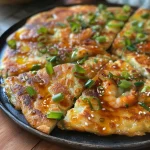 ['A close-up side view of a golden-brown Korean prawn pancake with visible prawns and green scallions.', 'Juicy Korean prawn pancake shown from the side, showcasing its crispy texture and vibrant ingredients.', 'A detailed view of a Korean prawn pancake, highlighting the shrimp and green onions embedded in the batter.', 'Side angle of a delicious Korean prawn pancake, emphasizing its crispy edges and moist interior.']