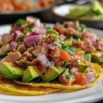 ['A close-up view of colorful tuna tostadas with avocado and toppings.', 'Side view of a delicious tuna tostada, showcasing fresh ingredients.', 'Juicy tuna tostadas stacked with avocado and cilantro on a plate.', 'Detail shot of vibrant tuna tostadas garnished with lime and onions.']