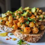 ['Close-up view of a slice of sourdough topped with spicy chickpeas and avocado.', 'Side view of a toasted bread slice with seasoned chickpeas and creamy avocado.', 'Juicy chickpea avocado toast garnished with chili flakes on a wooden surface.', 'Vibrant avocado toast with spicy chickpeas presented on a rustic plate.']