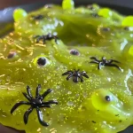 A side view close-up of vibrant green Halloween slime with glitter, containing googly eyes and small plastic spiders.
