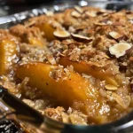 Close-up view of a golden-brown peach cobbler topped with oats and sliced almonds.